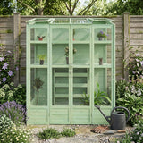 Victorian-style greenhouse in soft green, featuring glass panels, wooden shelves, and surrounded by vibrant flowers and plants