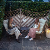 Two women enjoy drinks in a cozy grey and cream bistro set, surrounded by lush greenery and a charming wooden fence