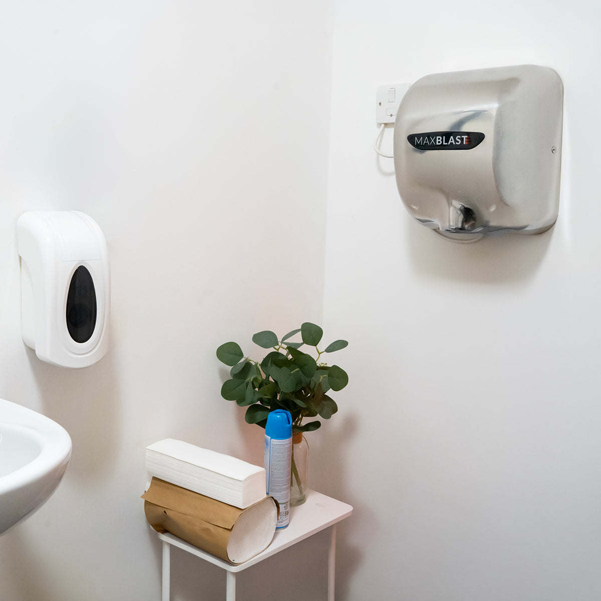 Sleek silver MAXBLAST hand dryer mounted on a white wall, beside a soap dispenser and a small potted plant on a table