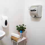 Sleek silver MAXBLAST hand dryer mounted on a white wall, beside a soap dispenser and a small potted plant on a table
