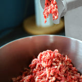 Red ground meat being extruded from the stainless steel KuKoo Commercial Meat Grinder into a large metal bowl, showcasing texture and freshness