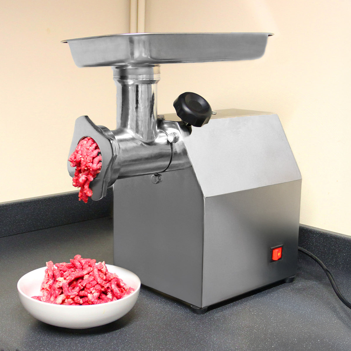 Stainless steel commercial meat grinder with ground meat extruding from the chute, accompanied by a bowl of minced meat on a black countertop