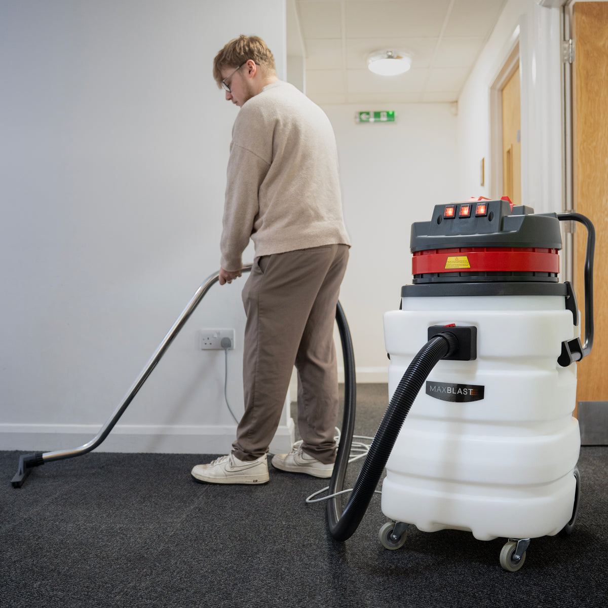 A person using a 90L Maxblast industrial vacuum cleaner on a carpeted floor, featuring a white tank and red accents, with a metal hose