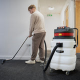 A person using a 90L Maxblast industrial vacuum cleaner on a carpeted floor, featuring a white tank and red accents, with a metal hose