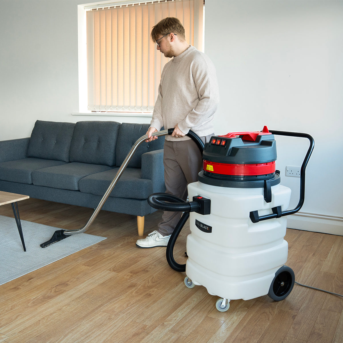 White 90L Maxblast industrial vacuum with red accents, being used on wooden floor near a gray sofa, featuring a long hose and nozzle