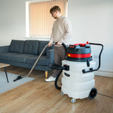 White 90L Maxblast industrial vacuum with red accents, being used on wooden floor near a gray sofa, featuring a long hose and nozzle