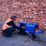 Electric log splitter in blue, with a person in an orange safety vest operating it, splitting a log against a brick wall background