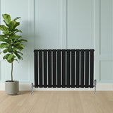 Flat black single column radiator with sleek vertical slats, complemented by chrome valves, positioned beside a potted plant in a modern room