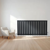 Flat black single column radiator with sleek vertical slats, paired with chrome valves, positioned beside a modern armchair in a bright room