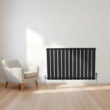 Flat black single column radiator with vertical slats, positioned beside a modern white armchair in a bright room with wooden flooring