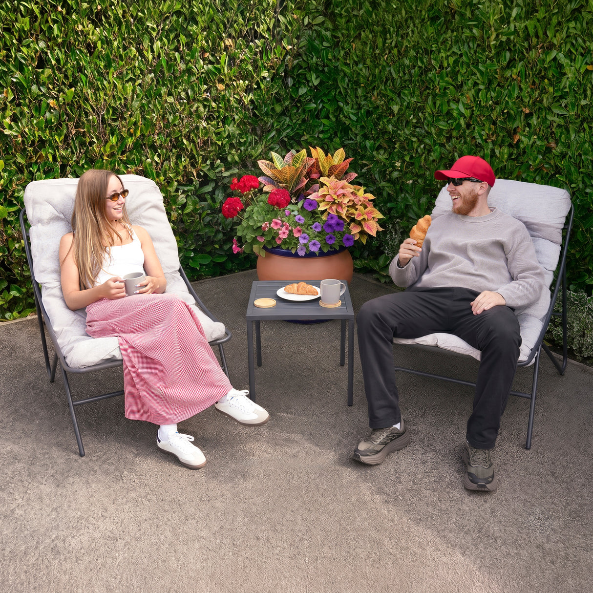 Stylish grey 3 piece bistro set featuring two cushioned chairs and a small table, set against vibrant greenery and colorful flowers