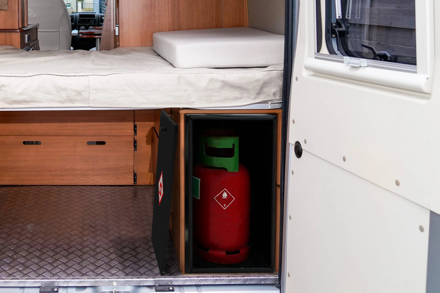Red gas cylinder stored in a black cabinet beneath a wooden bed in a camper van, with a light-colored mattress visible above