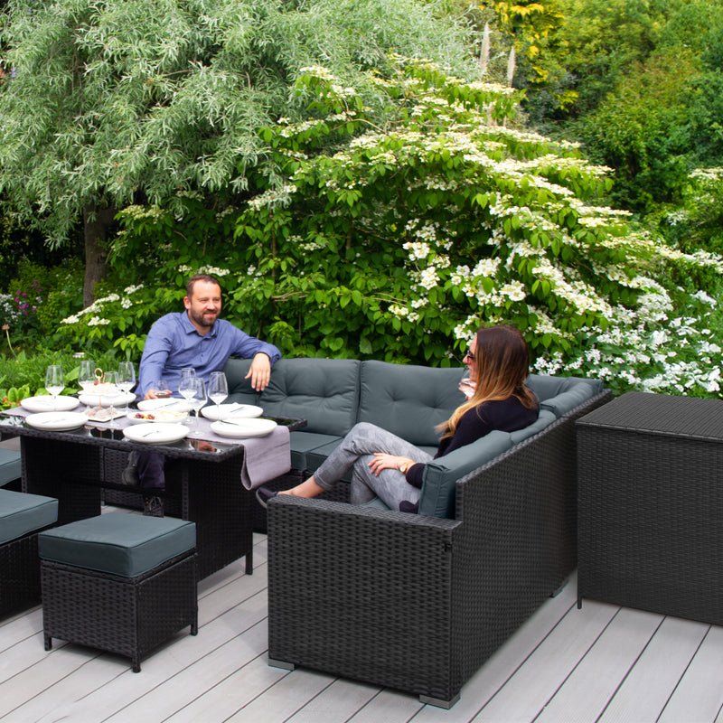 Stylish outdoor seating set with dark wicker furniture, gray cushions, and a dining table, surrounded by lush greenery and flowers