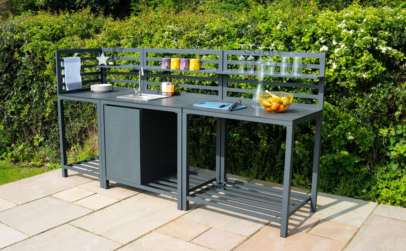 Sleek black outdoor kitchen island with slatted back, featuring a sink, countertop, and decorative fruit bowl, set in a lush garden
