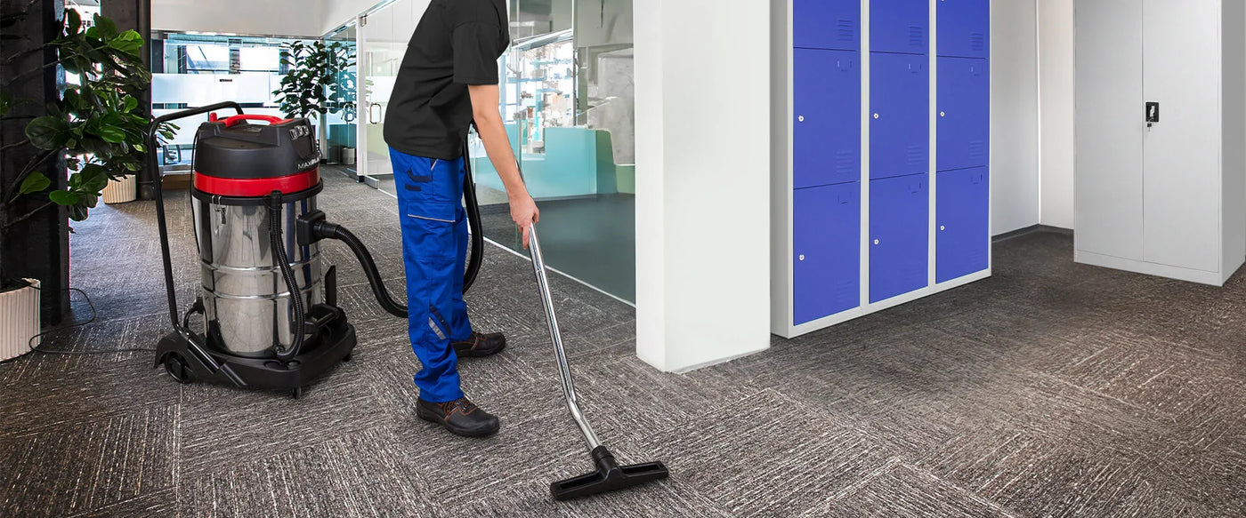 Person using a vacuum cleaner in an office setting with blue lockers and a carpeted floor.