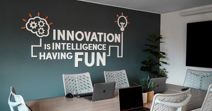 A modern conference room featuring a dark gray wall with the text "Innovation is Intelligence Having Fun," surrounded by plants and sleek furniture
