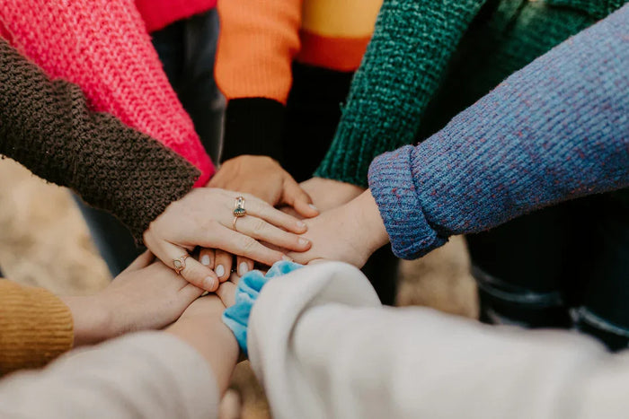 A diverse group of hands stacked together, showcasing colorful knitted sweaters in shades of pink, green, and blue, symbolizing unity