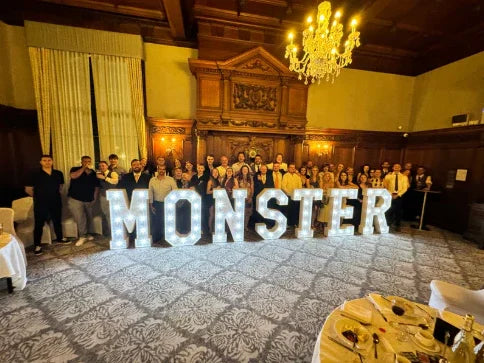 A large illuminated sign spelling "MONSTER" stands in a grand room filled with people dressed in formal attire, showcasing elegant decor and a chandelier